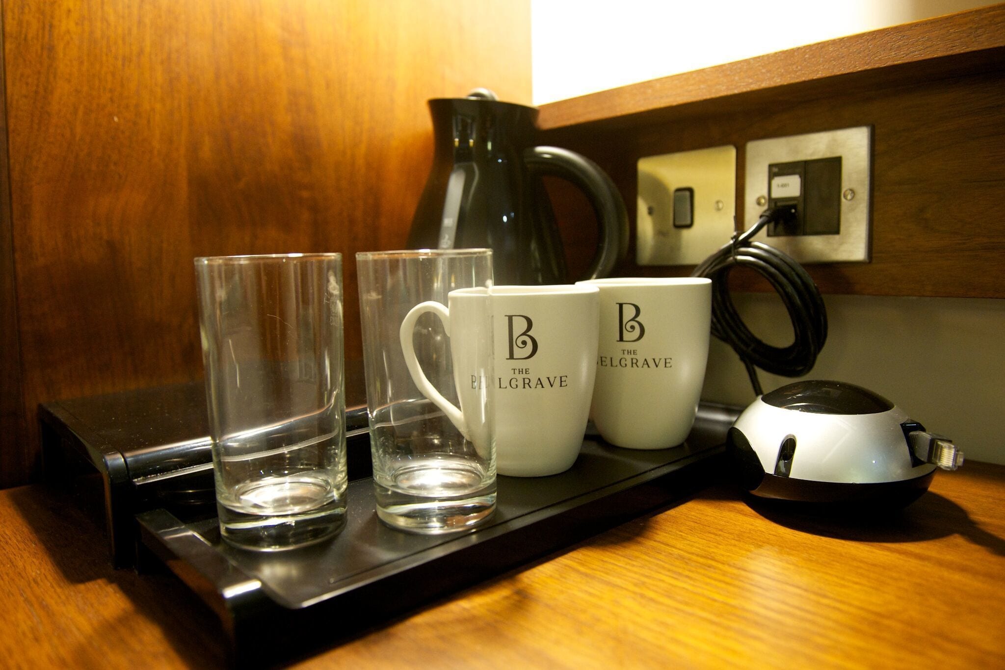 A tray with two glasses and two mugs on a wooden surface, a black electric kettle, and a power outlet.