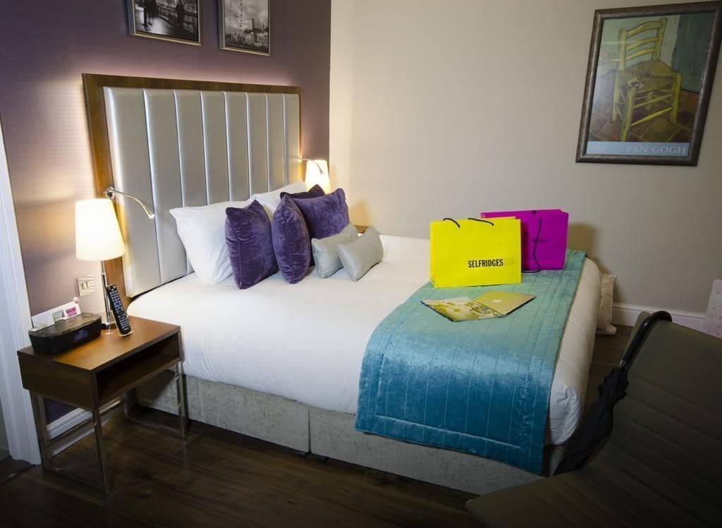A hotel room with a bed, a nightstand, a lamp, a chair, and a painting on the wall.