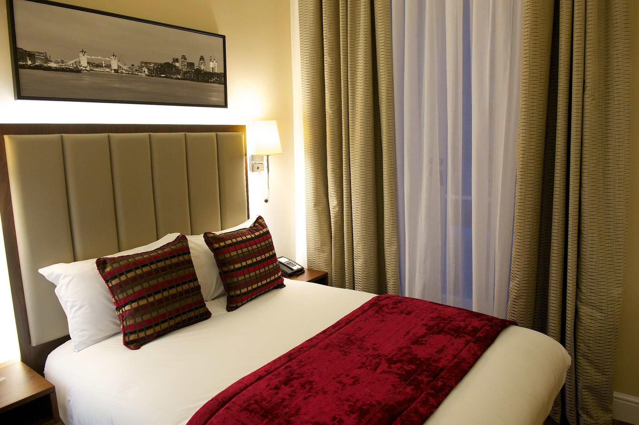 A hotel room with a large bed, two pillows, a red blanket, a nightstand with a lamp, and a framed picture on the wall.