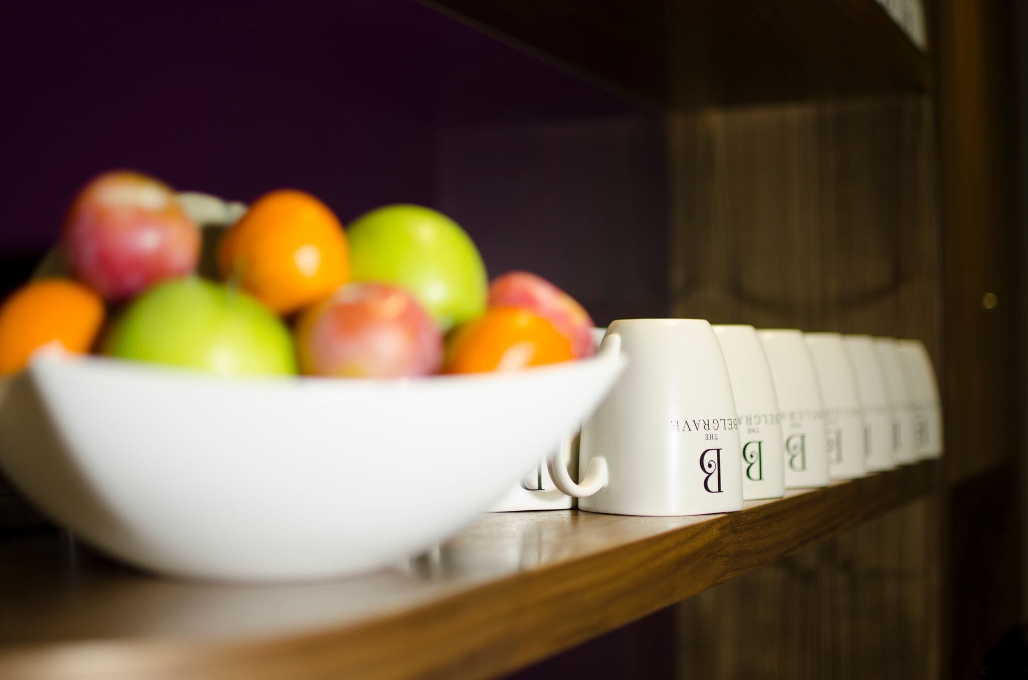 A white bowl filled with colorful fruits on a wooden surface, next to a row of white mugs with a logo.