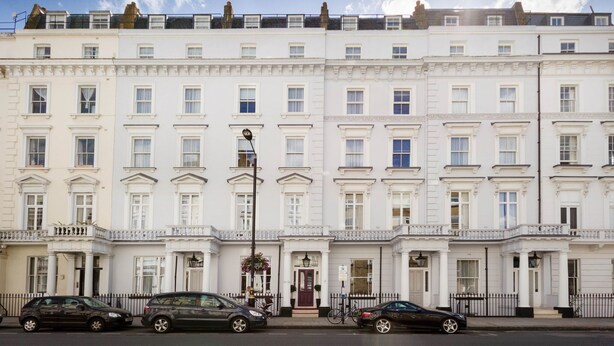 A row of white townhouses with uniform windows and balconies, a street in front, and parked cars. - The Belgrave (London)