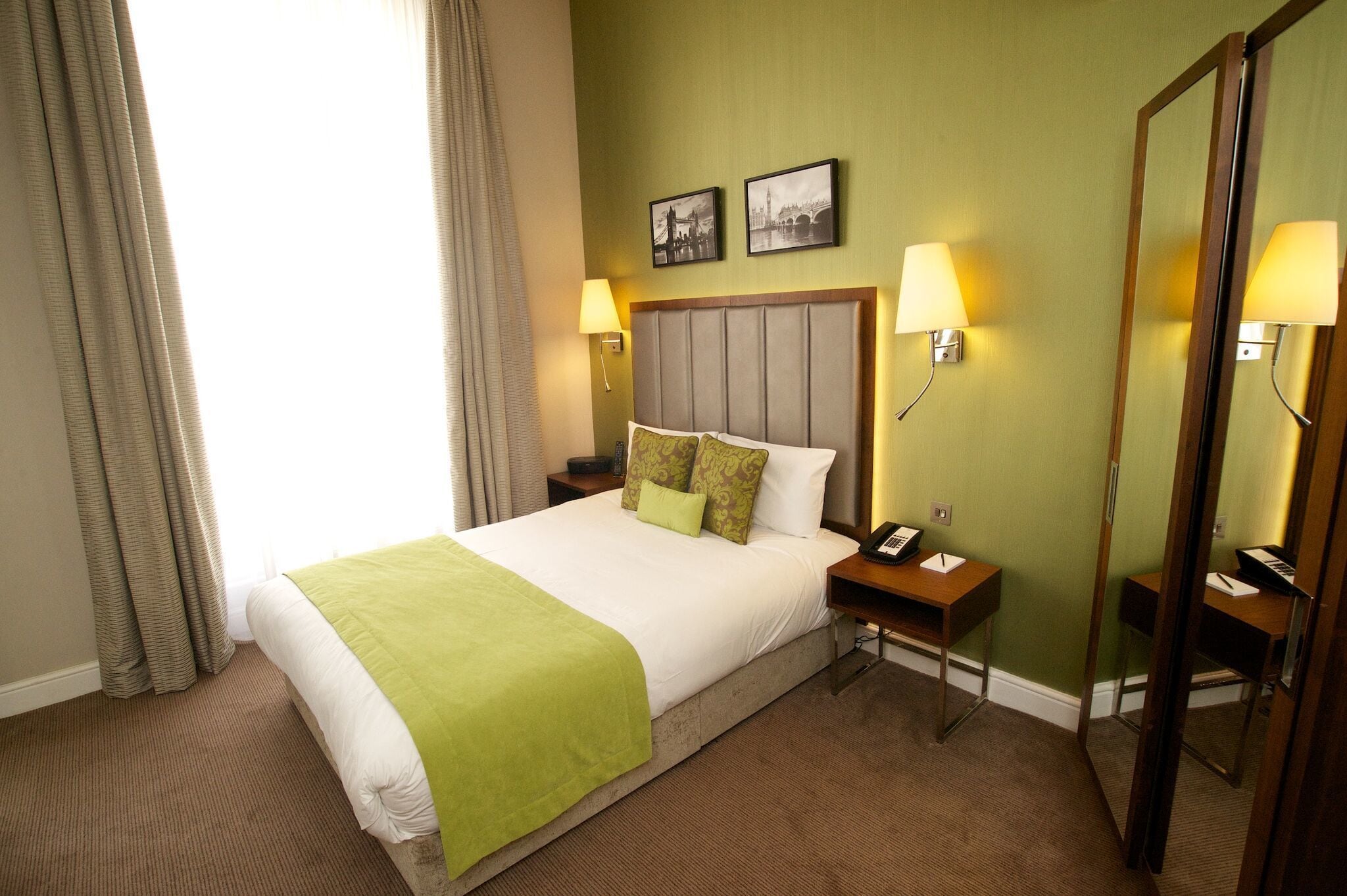 A hotel room with a bed, two bedside tables, a nightstand, a mirror, and two wall-mounted lamps.