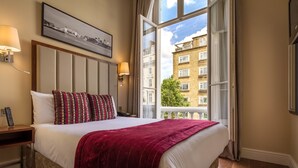 A hotel room with a large bed, a nightstand, a lamp, a framed picture on the wall, and a window with a view of buildings. - The Belgrave (London)