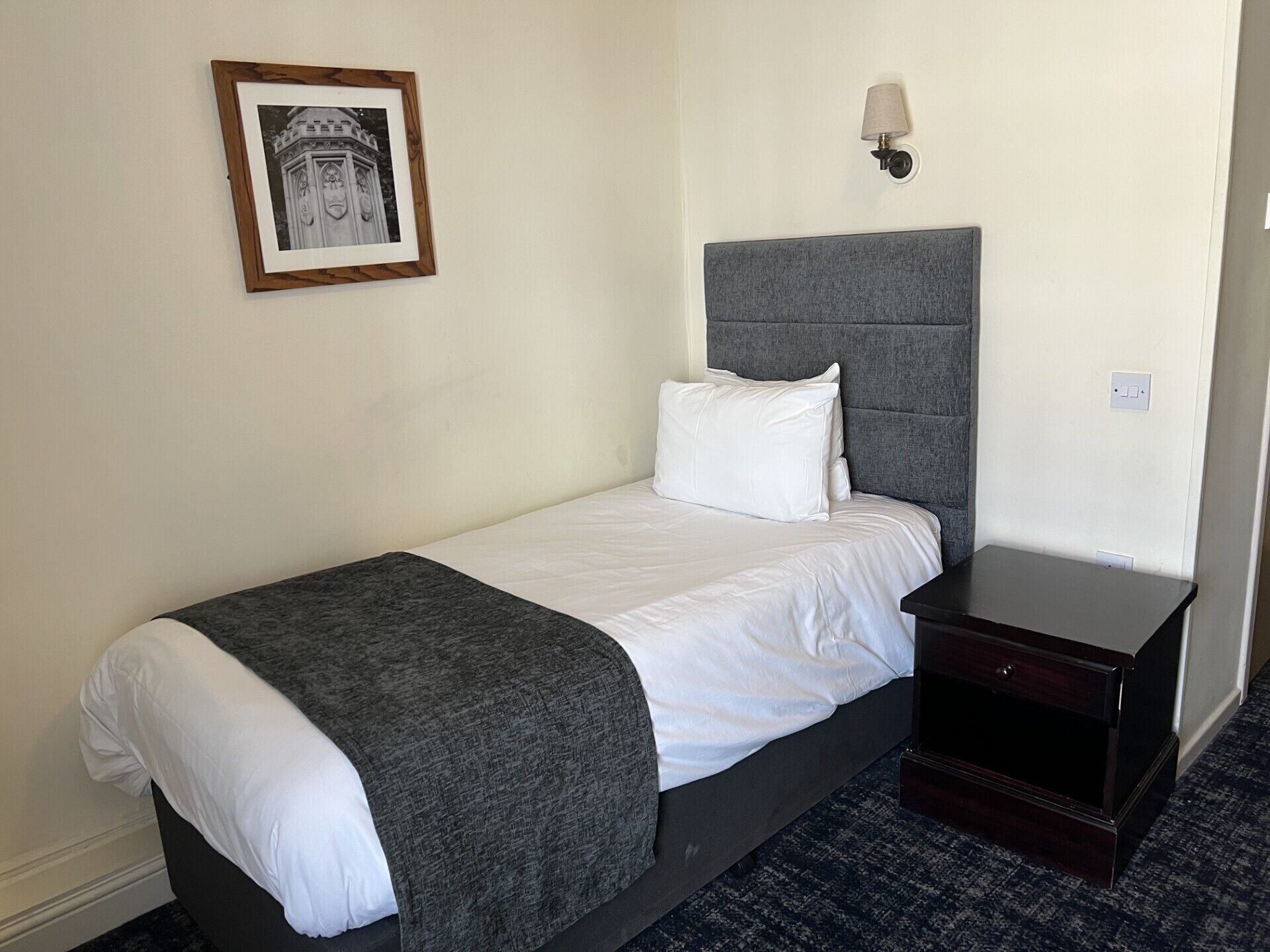 Una cama individual con cabecera gris, almohada blanca, manta gris y una mesita de noche pequeña. En la pared, un cuadro enmarco.