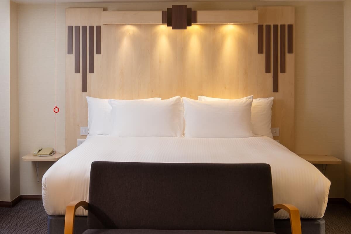 A hotel room with a large bed, a bedside table with a lamp, a chair, and a wall-mounted phone.