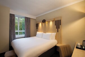 Ein Hotelzimmer mit einem großen Bett, einem Nachttisch mit Kaffeemaschine und Blick auf Bäume durch das Fenster.