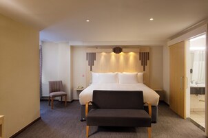 A hotel room with a large bed, a bedside table, a chair, and a bench.