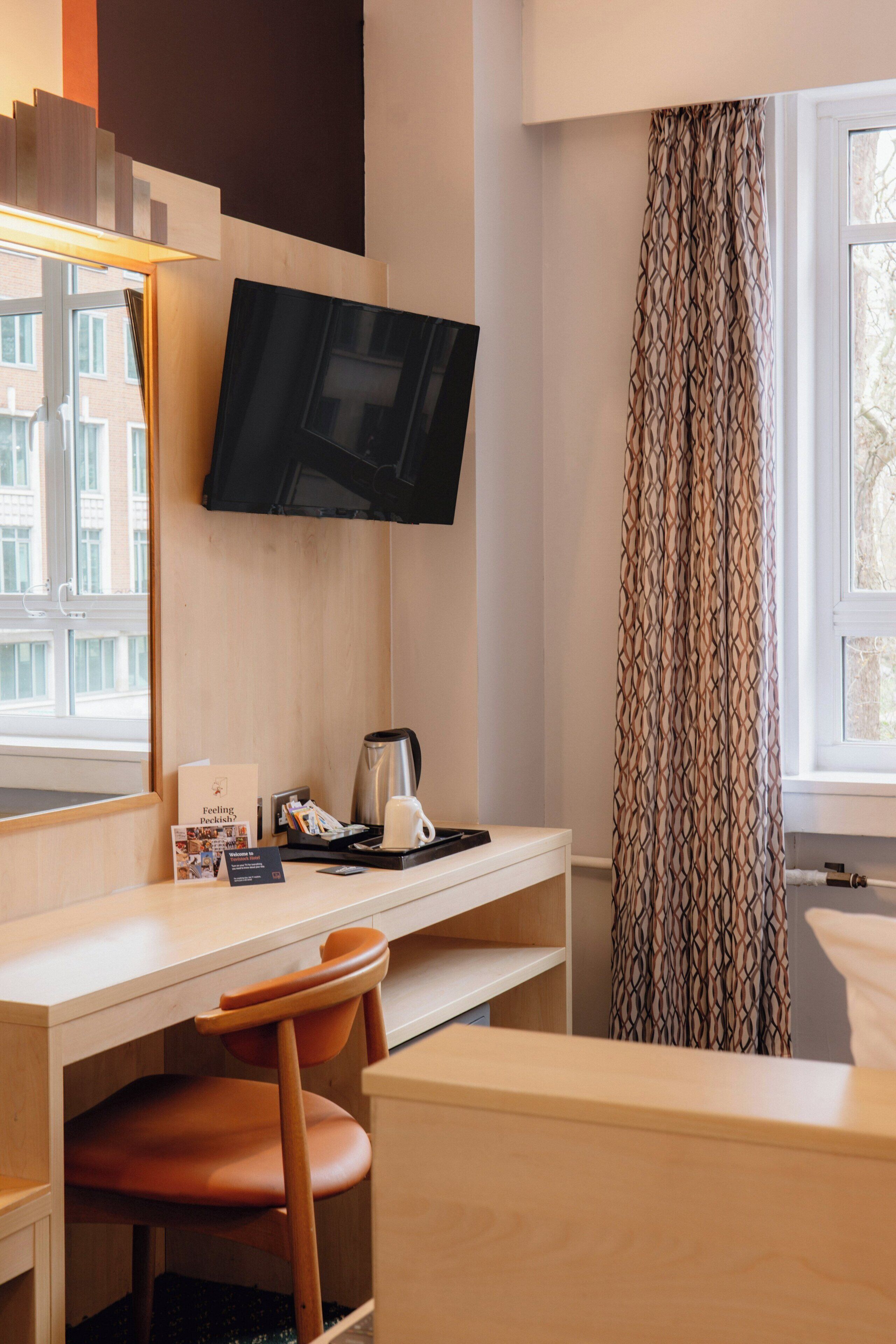 A hotel room with a wooden desk, a television mounted on the wall, a chair, a window with patterned curtains, and a view of buildings outside.