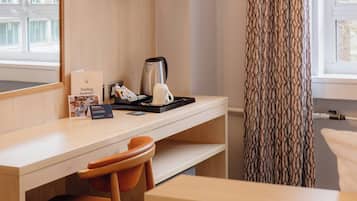 A hotel room with a wooden desk, a television mounted on the wall, a chair, a window with patterned curtains, and a view of buildings outside.