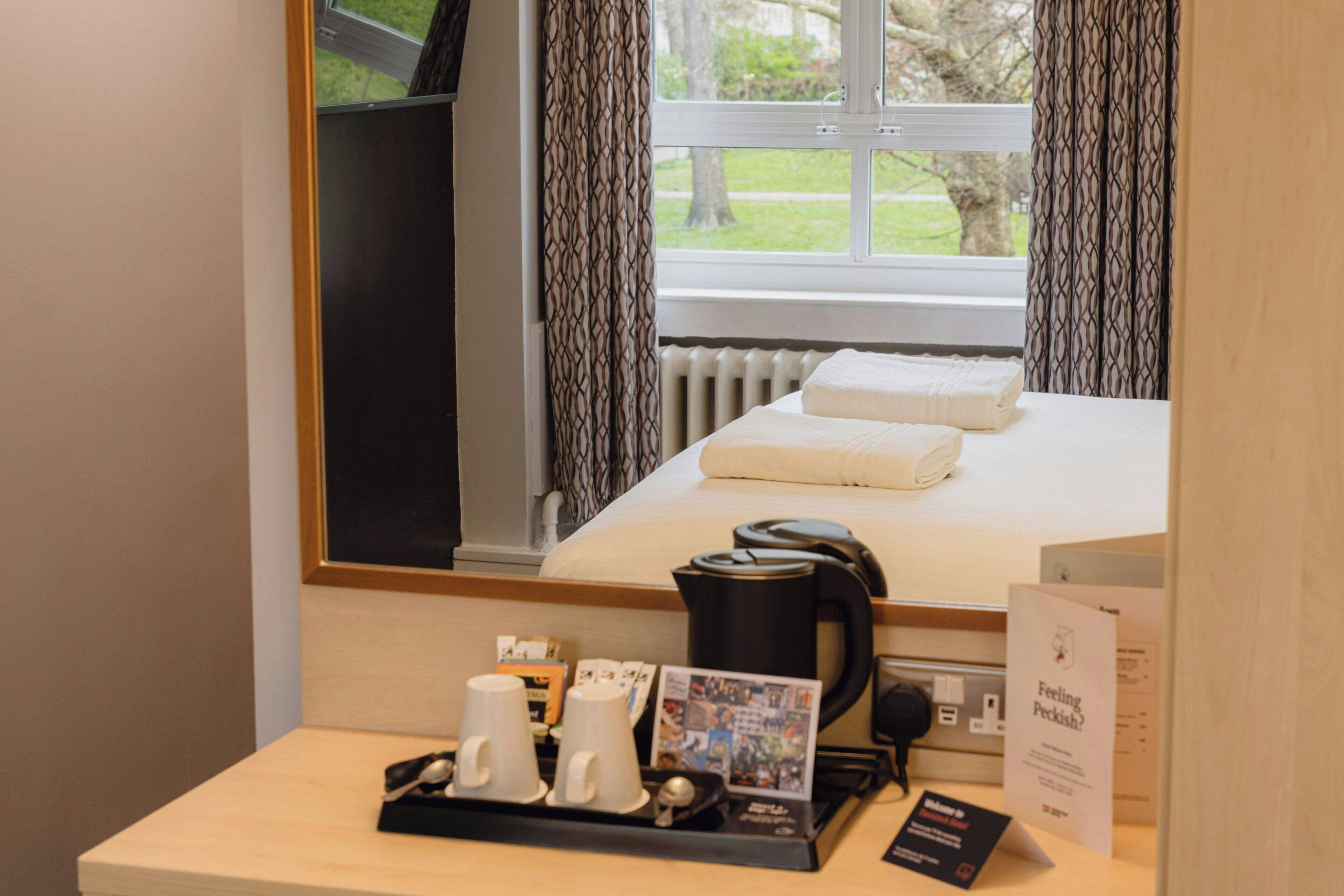 A hotel room with a bed, a coffee maker, a tray with mugs, a framed picture, and a brochure on the desk.