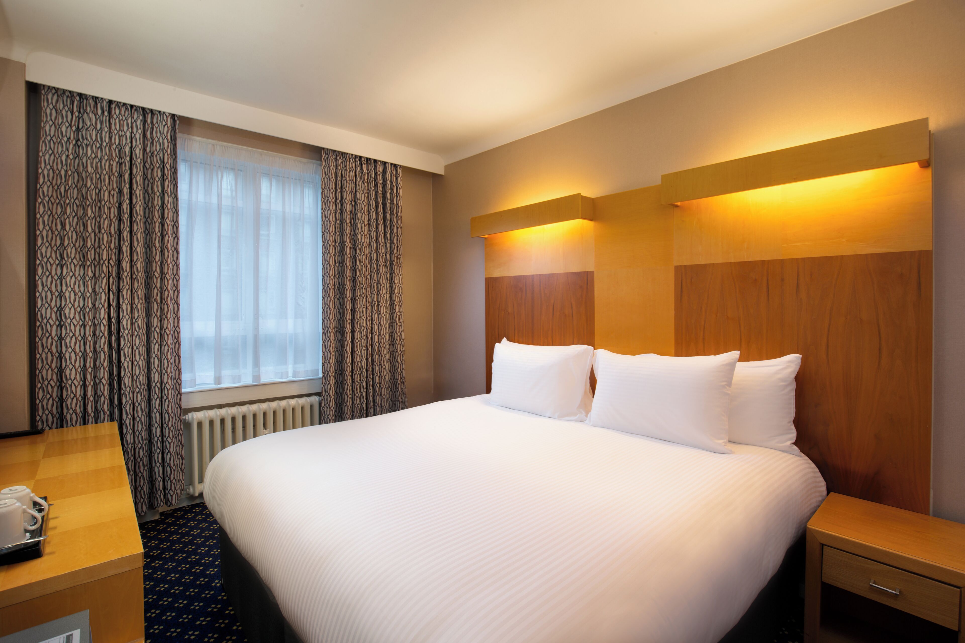 A hotel room with a large bed, a wooden headboard, a bedside table, a desk with a telephone, and a window with curtains.