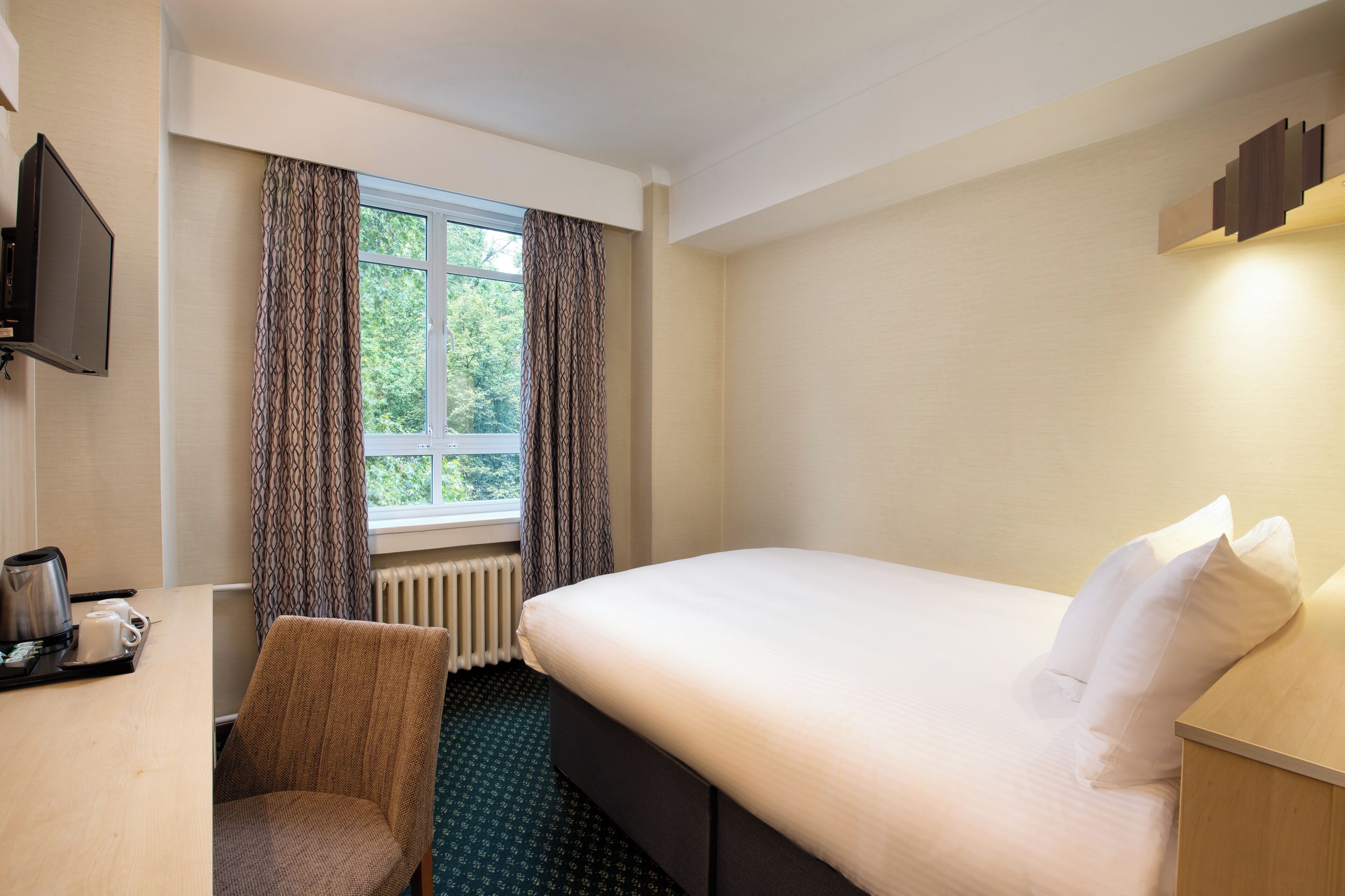 A hotel room with a bed, a desk with a kettle, a chair, and a window with curtains.