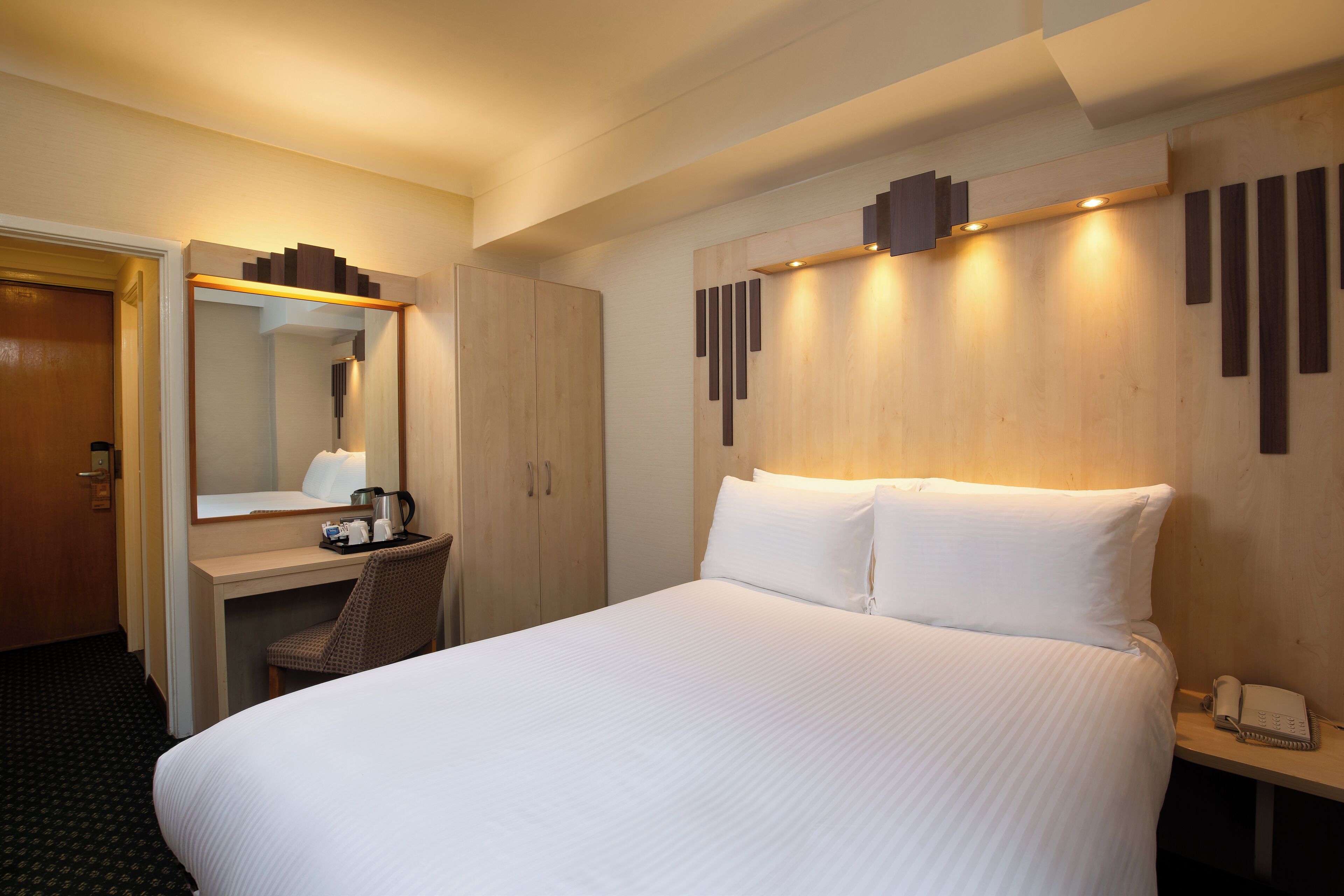 A hotel room with a large bed, a desk with a chair, a mirror, and a bedside table with a phone.