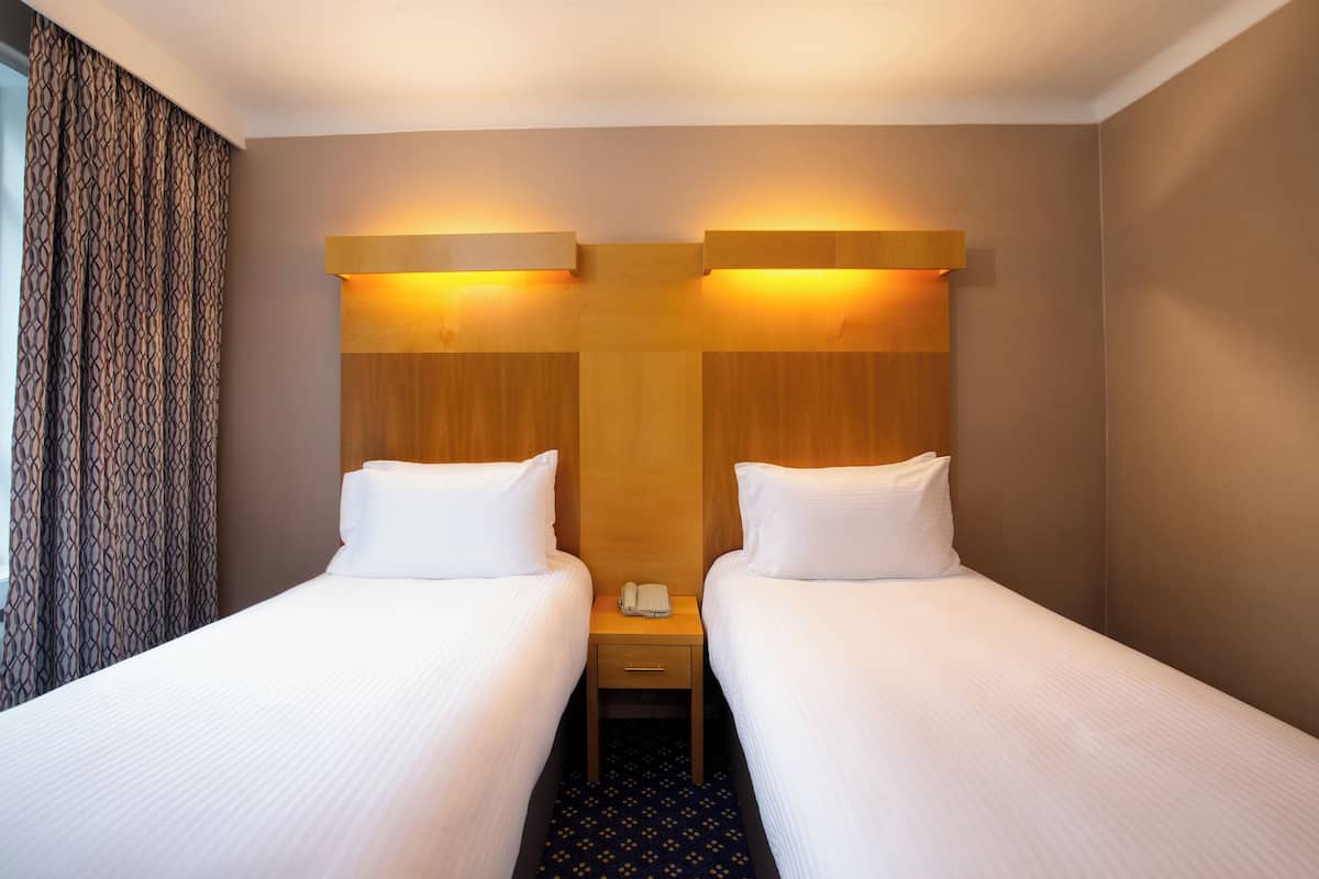 A hotel room with two single beds, a wooden headboard, a bedside table with a phone, and a patterned curtain.