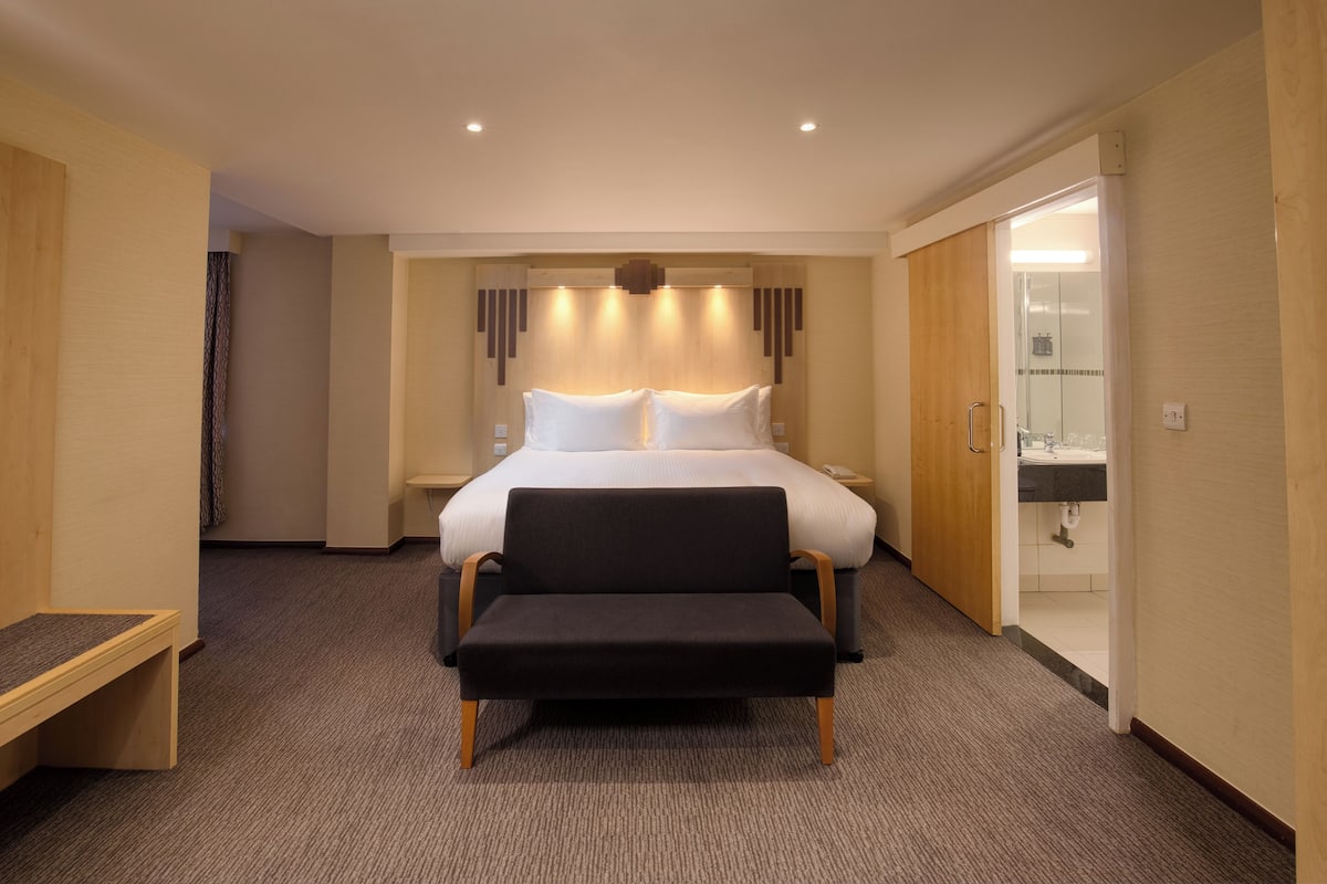 A hotel room with a large bed, a bench, a bedside table, and a bathroom visible through an open door.