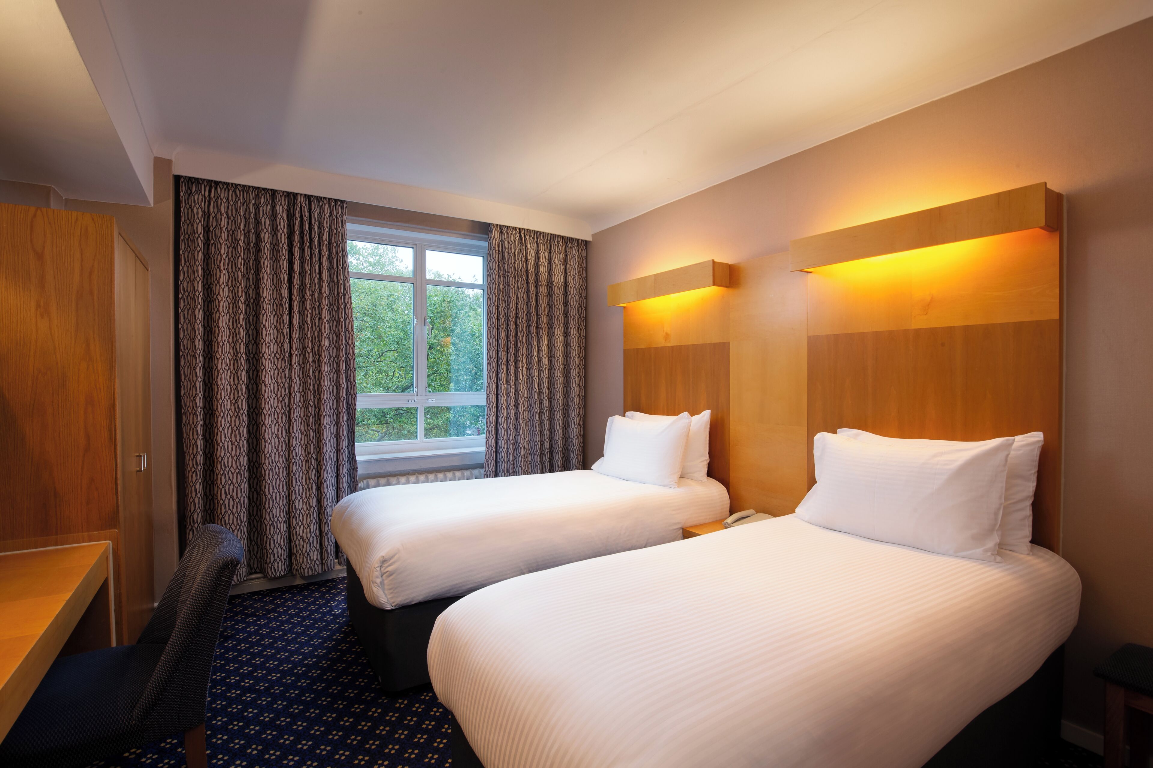 A hotel room with two beds, a desk, a chair, and a window with curtains.