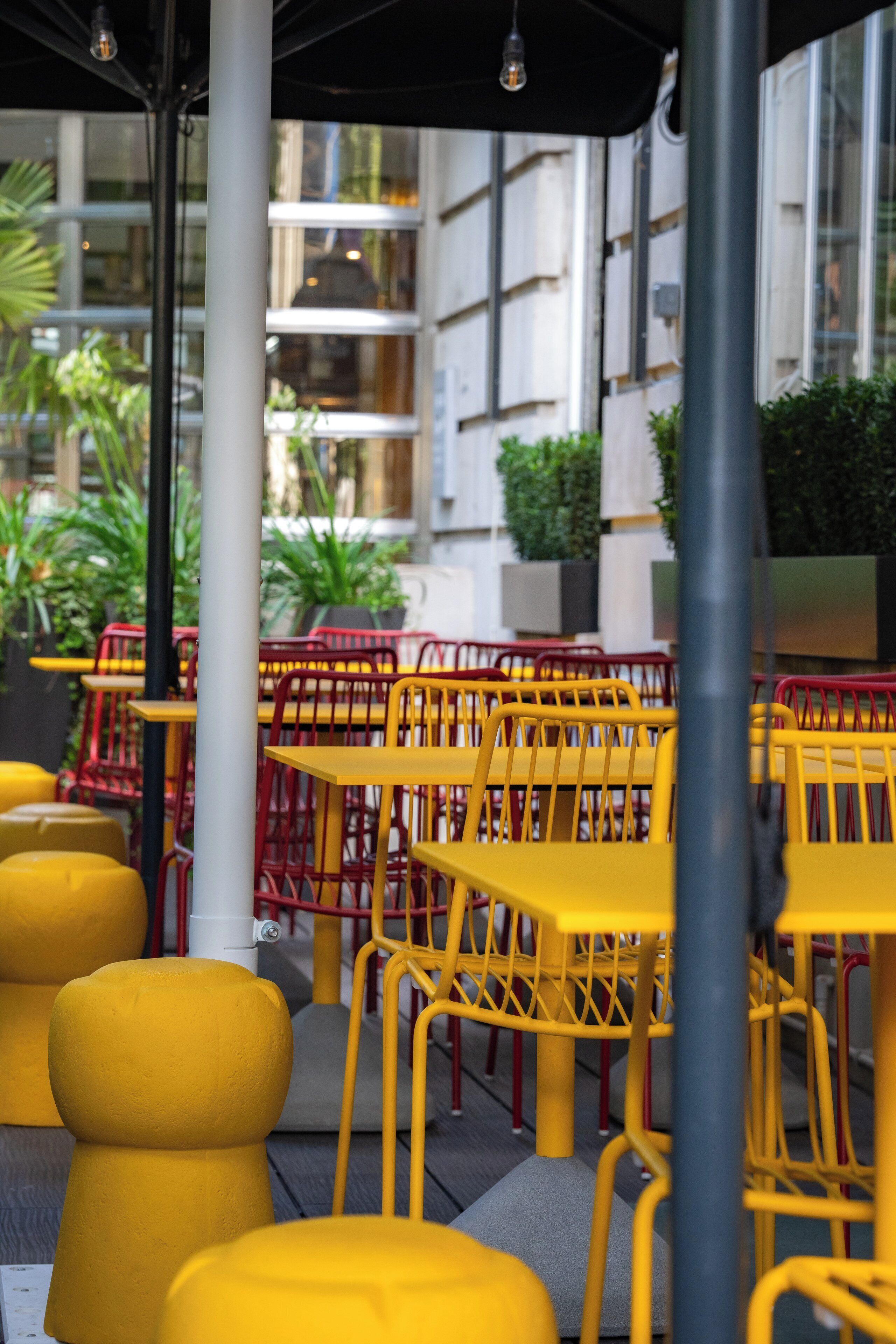 A seating area with yellow tables and chairs, surrounded by greenery and a modern building in the background.