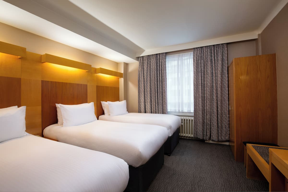 A hotel room with three single beds, a wooden wardrobe, a desk, and a window with patterned curtains.