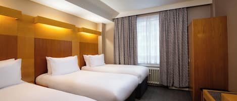 A hotel room with three single beds, a wooden wardrobe, a desk, and a window with patterned curtains.