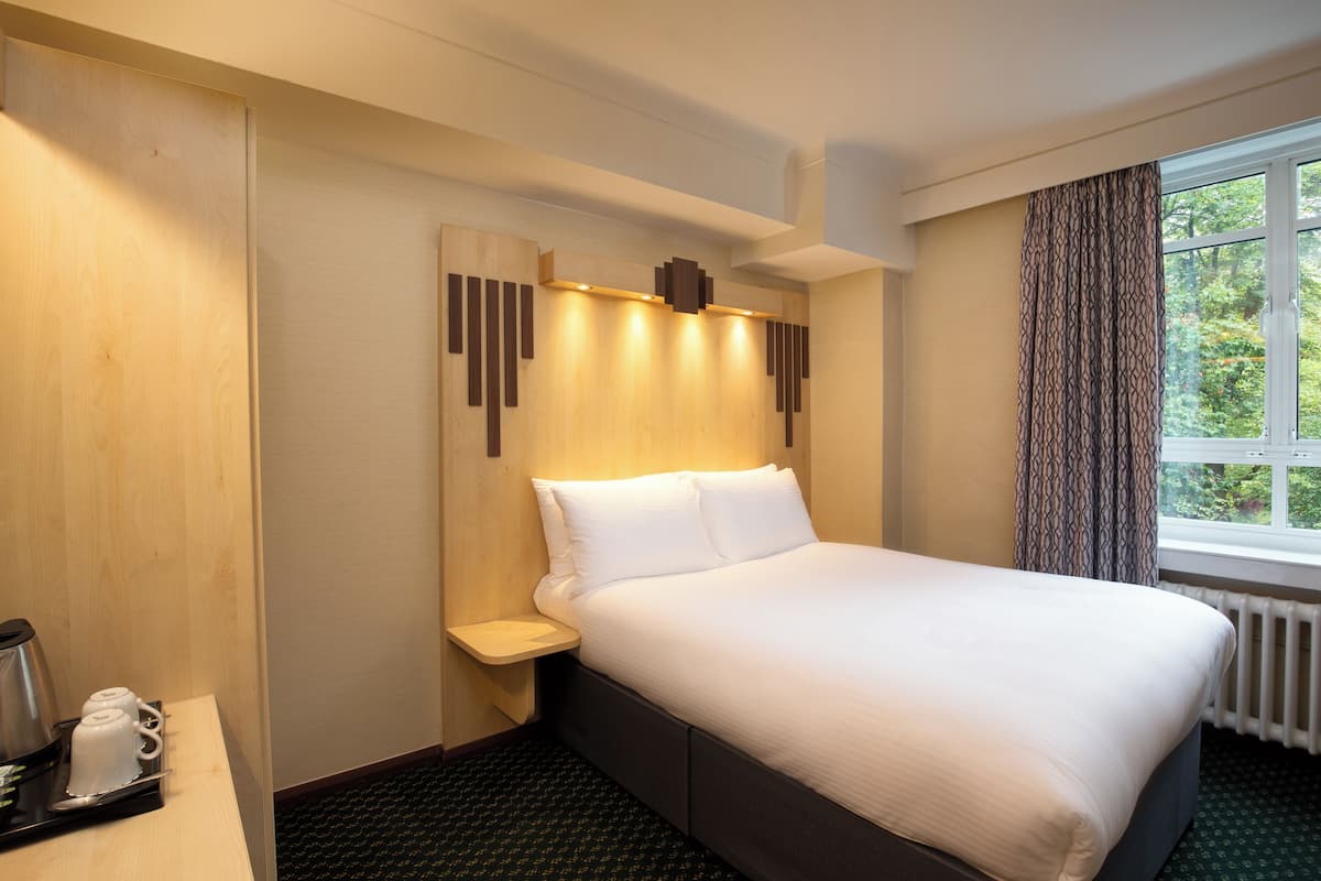 A hotel room with a large bed, a bedside table with a coffee maker, a window with curtains, and a wooden headboard.