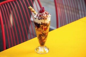 Un postre en un vaso sobre una mesa amarilla, con sillas rojas al fondo.