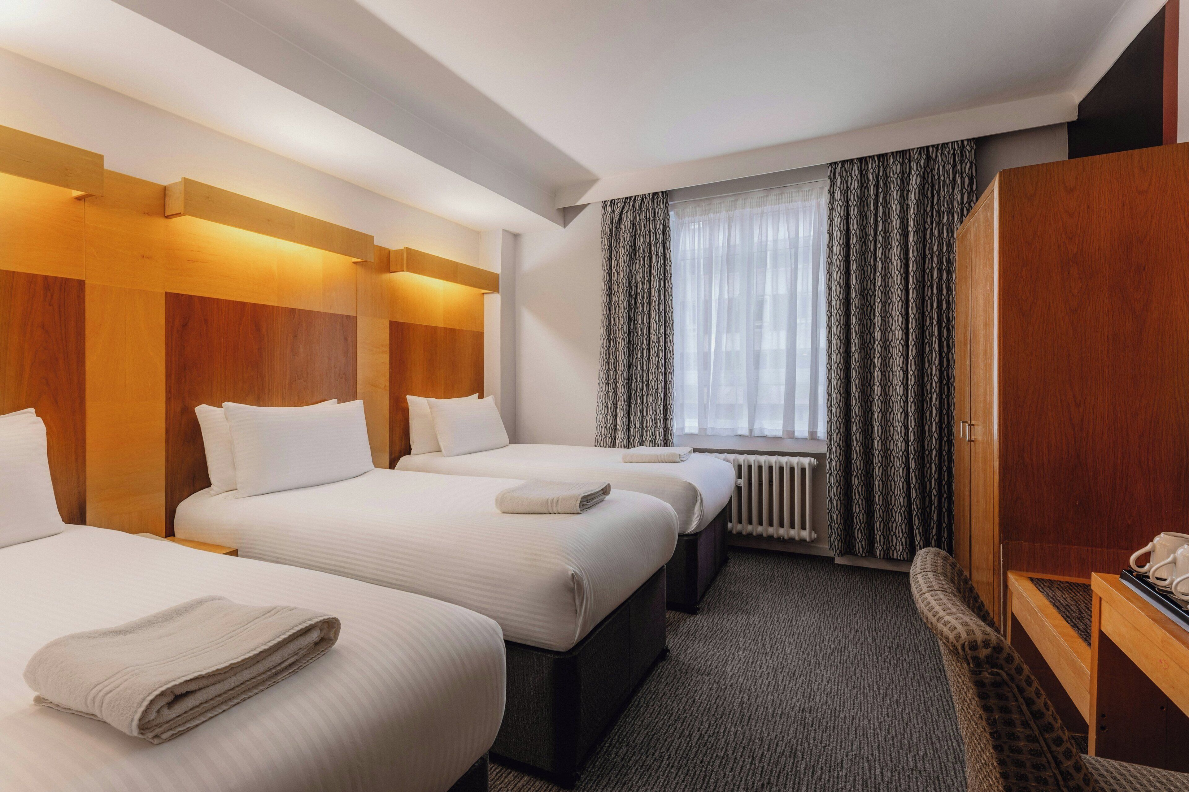 Habitación de hotel con dos camas, cabecero de madera, radiador, escritorio con teléfono y ventana con cortinas estampadas.