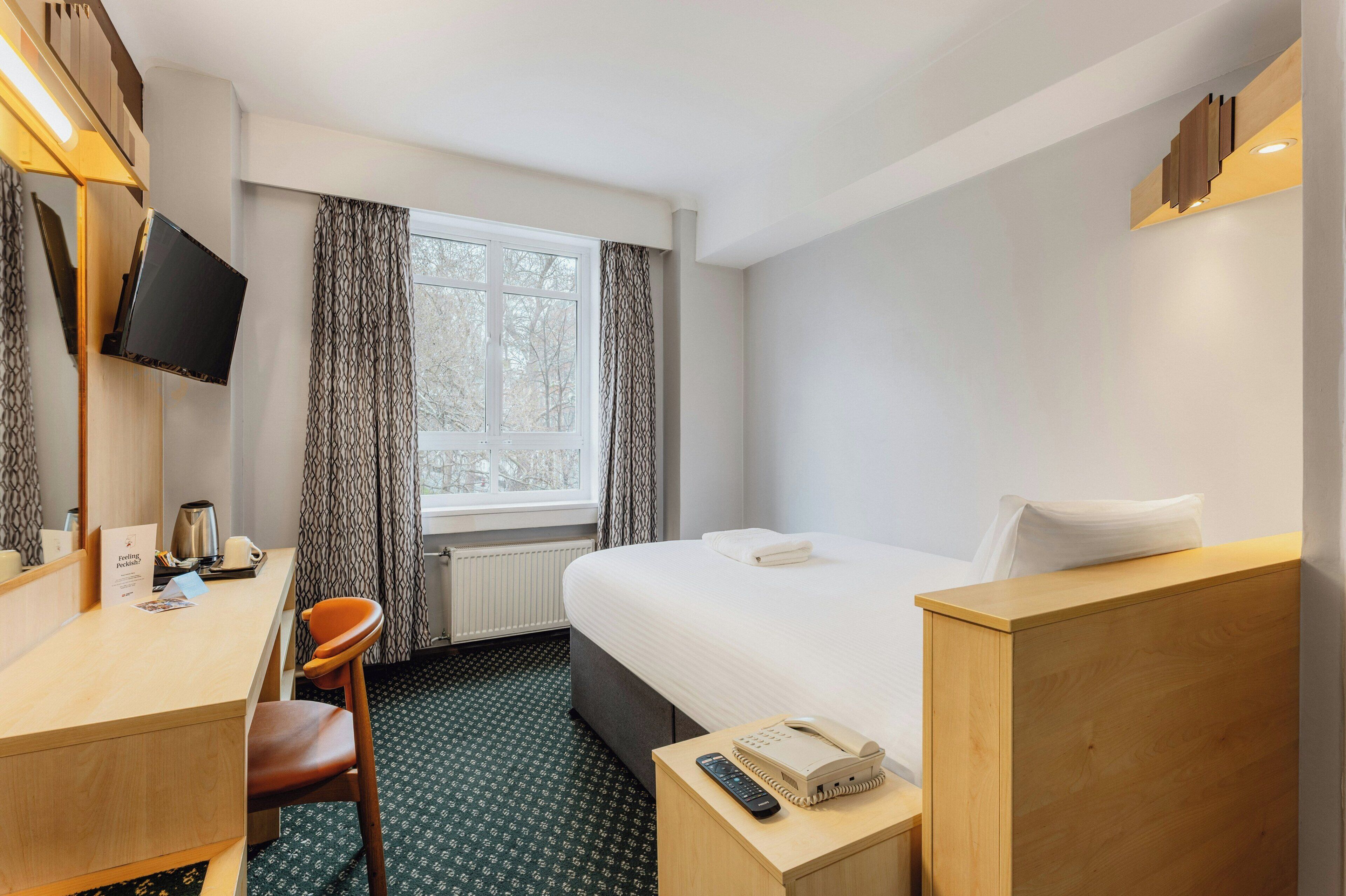 Una habitació d'hotel amb un llit, una taula, una cadira, un televisor muntat a la paret i una finestra amb cortines.