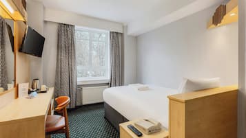 Una habitació d'hotel amb un llit, una taula, una cadira, un televisor muntat a la paret i una finestra amb cortines.