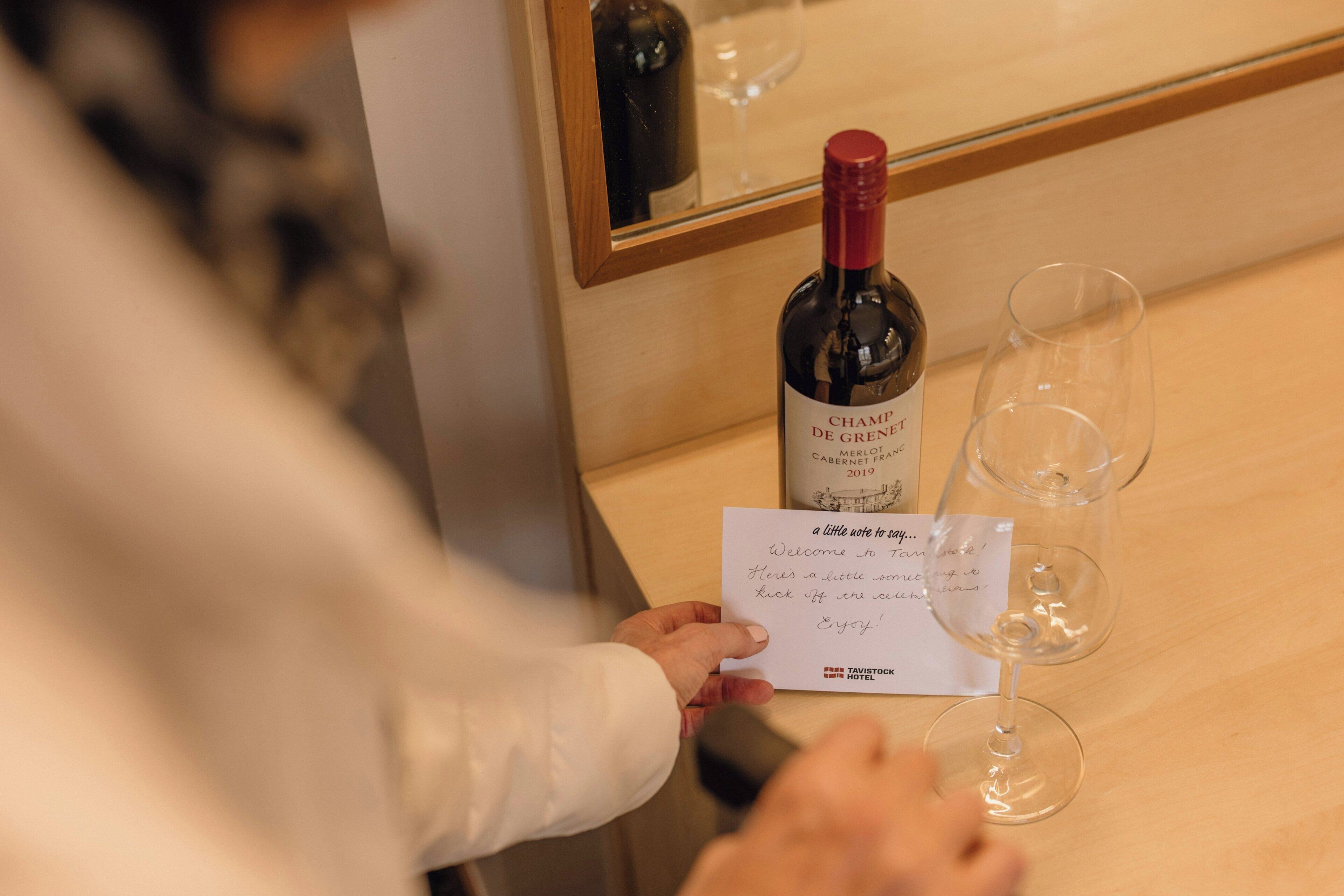 Une personne tenant une bouteille de vin et un verre, avec un petit mot à côté.