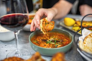 A bowl of curry with a hand lifting a piece of naan bread, a glass of red wine, and a plate with lemon slices on a table.