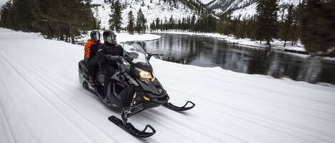 Un vehículo para nieve en un camino cubierto de nieve, con un lago y montañas al fondo.