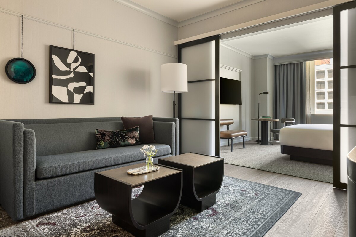 Une chambre d’hôtel moderne avec un canapé gris, deux tables d’appoint noires, un grand miroir et une vue sur la chambre à coucher.