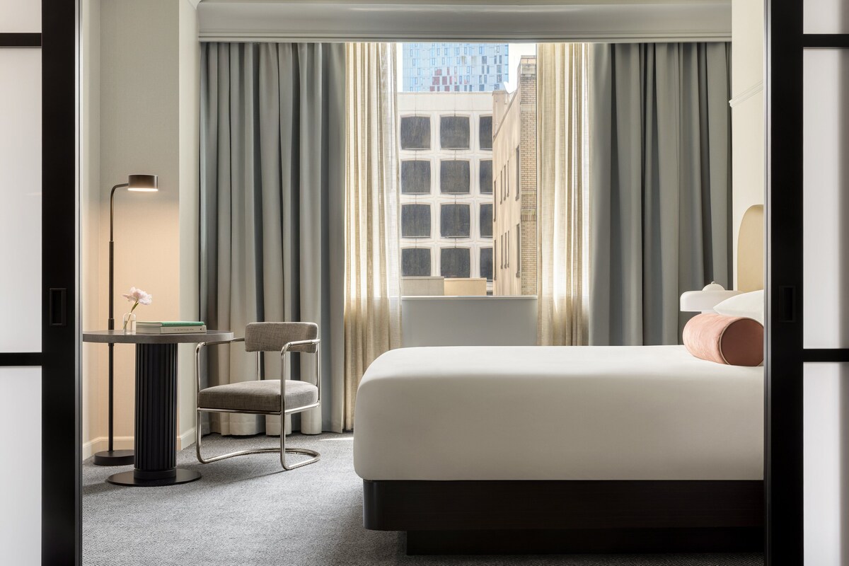 Une chambre d’hôtel avec un lit, un bureau et une chaise, une lampe, et une vue sur les bâtiments par la fenêtre.