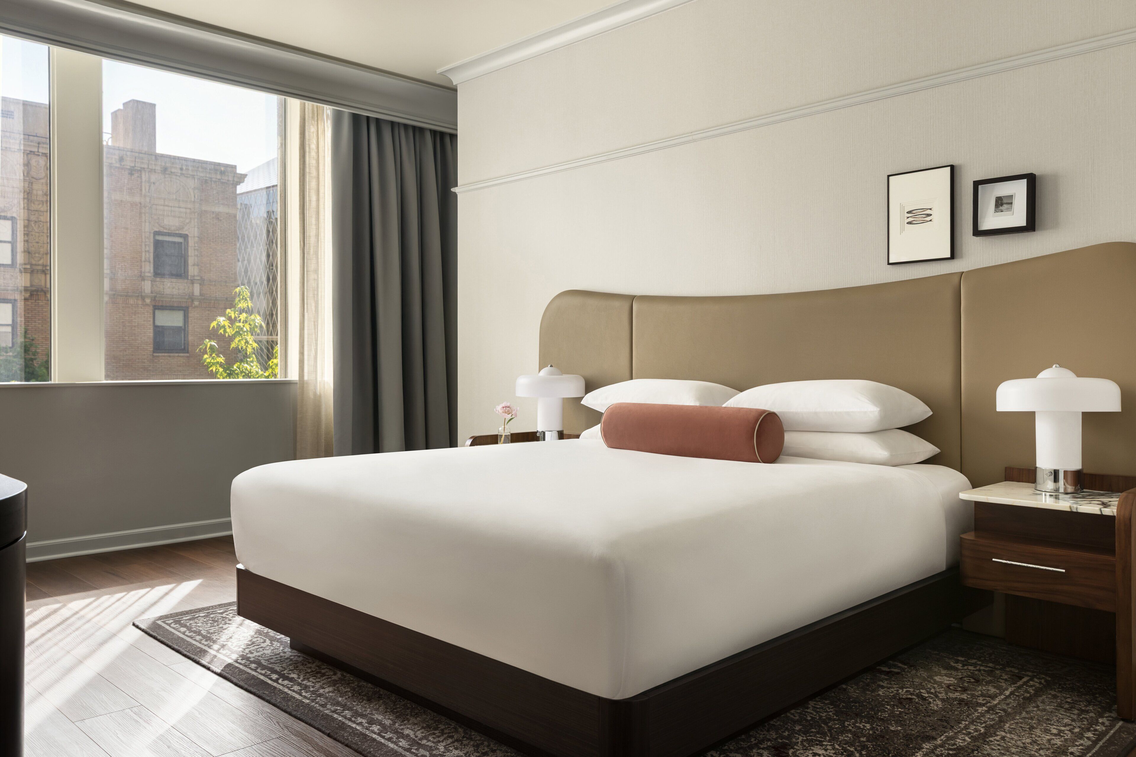Habitación de hotel con una cama grande, una mesita de noche con lámpara, una ventana con cortinas y vistas a edificios.