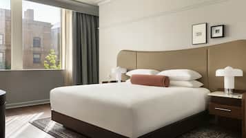Habitación de hotel con una cama grande, una mesita de noche con lámpara, una ventana con cortinas y vistas a edificios.