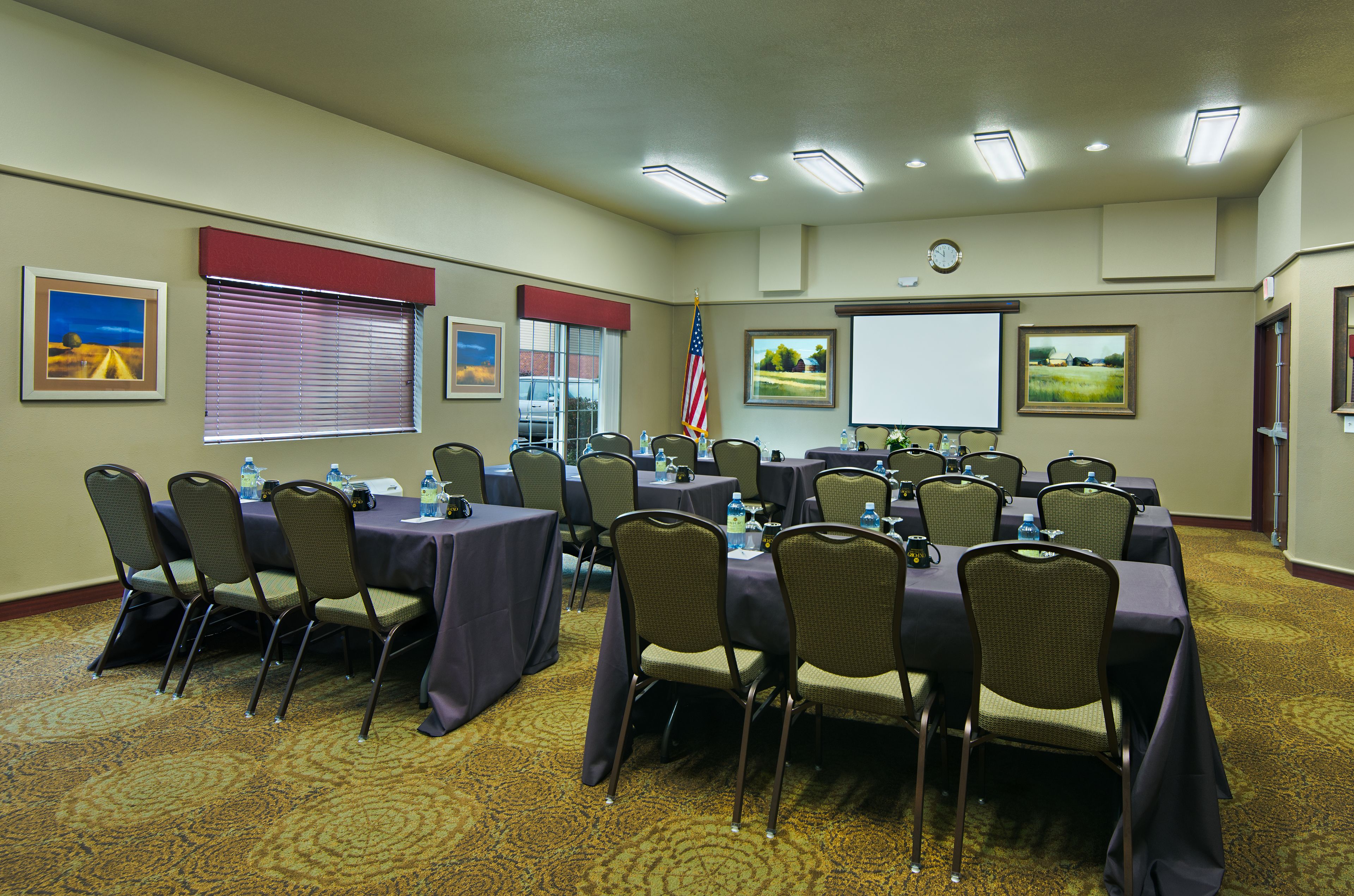 A conference room with multiple tables and chairs, a projector screen, and an American flag.
