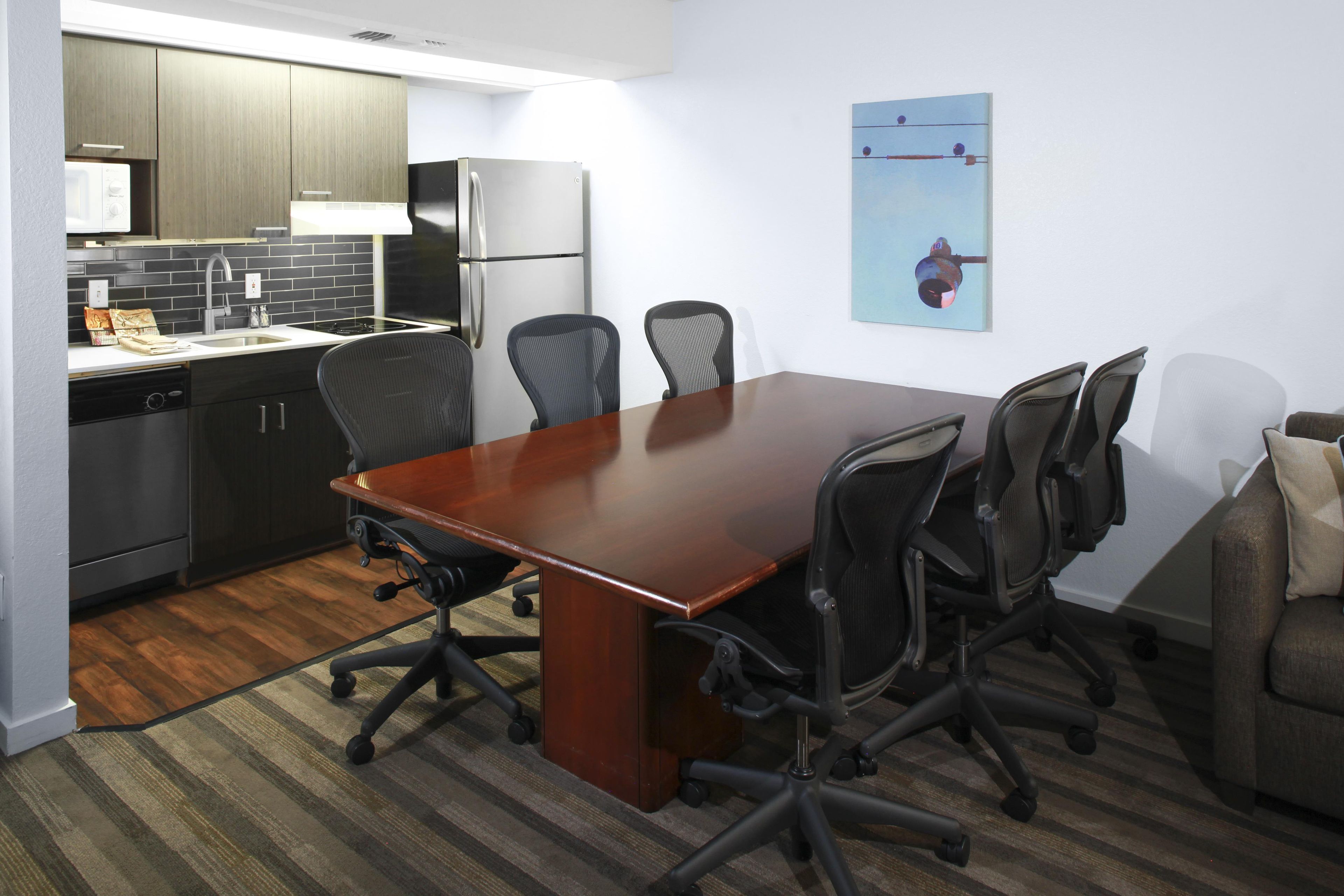 Uma sala de conferências com uma mesa comprida, seis cadeiras e uma área de cozinha com geladeira, micro-ondas e armários.