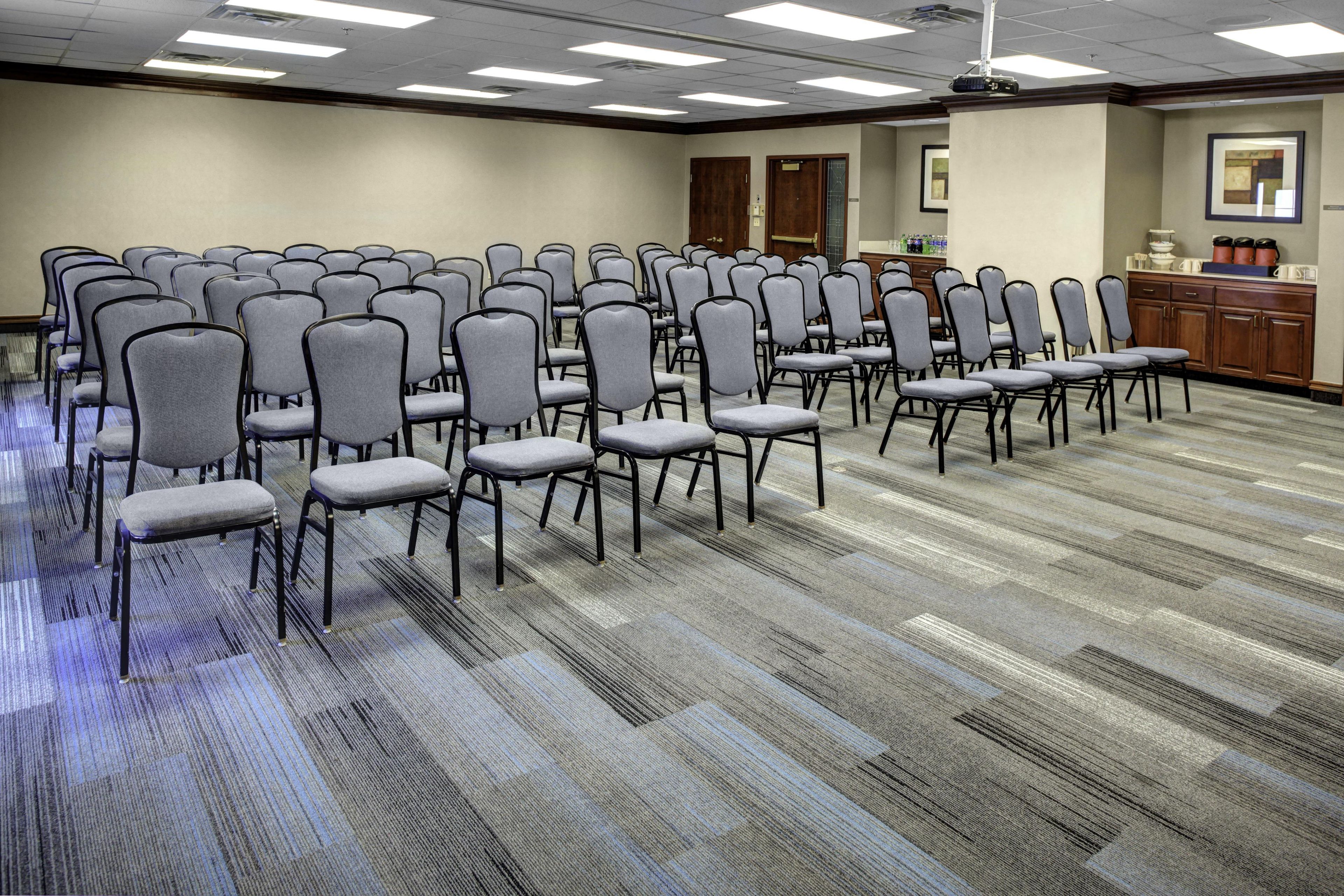 Une salle de conférence avec des rangées de chaises grises, un sol recouvert de moquette et un coin cuisine.