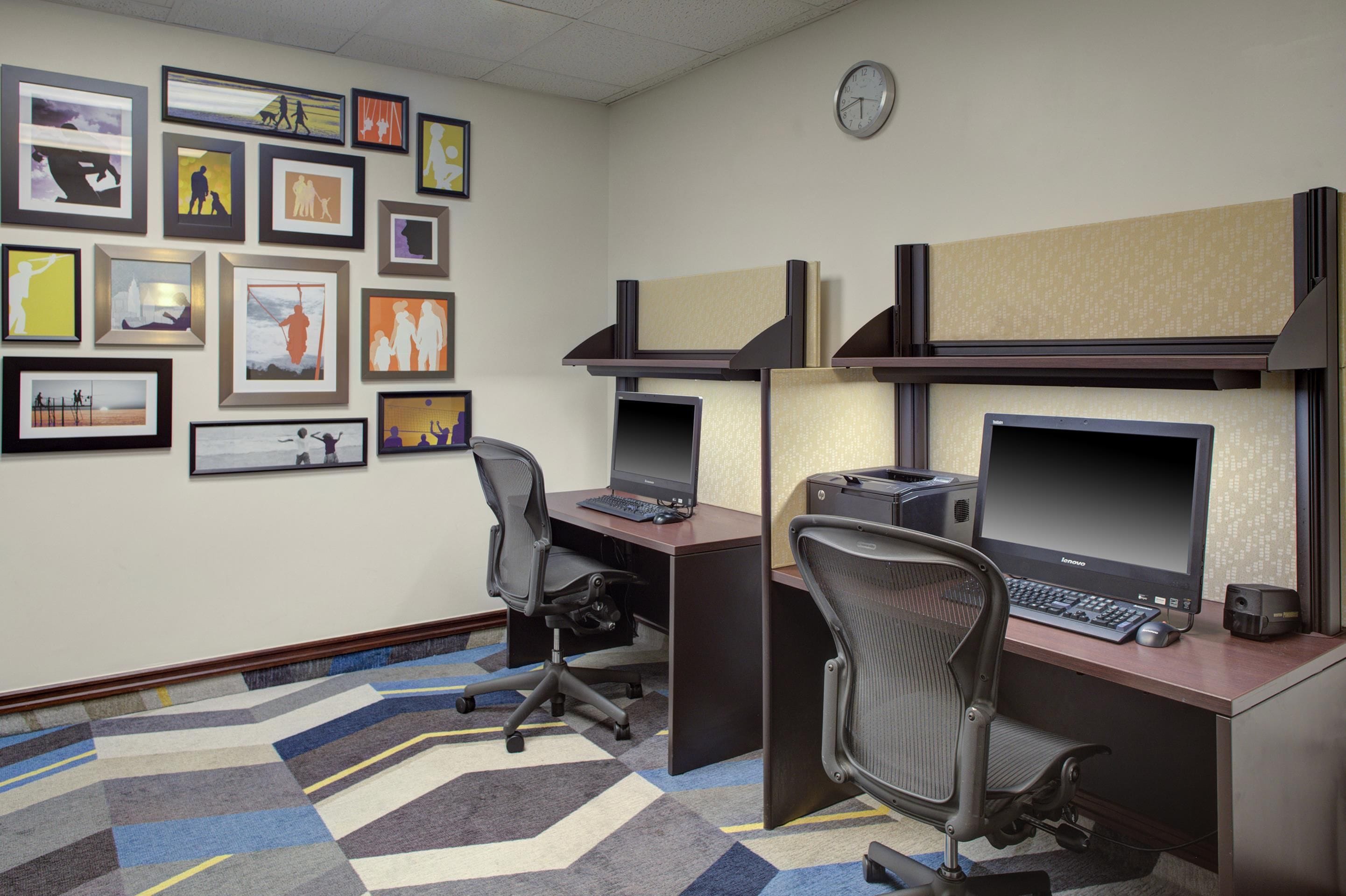 A cubicle office with multiple monitors, a clock, and framed artwork on the wall.