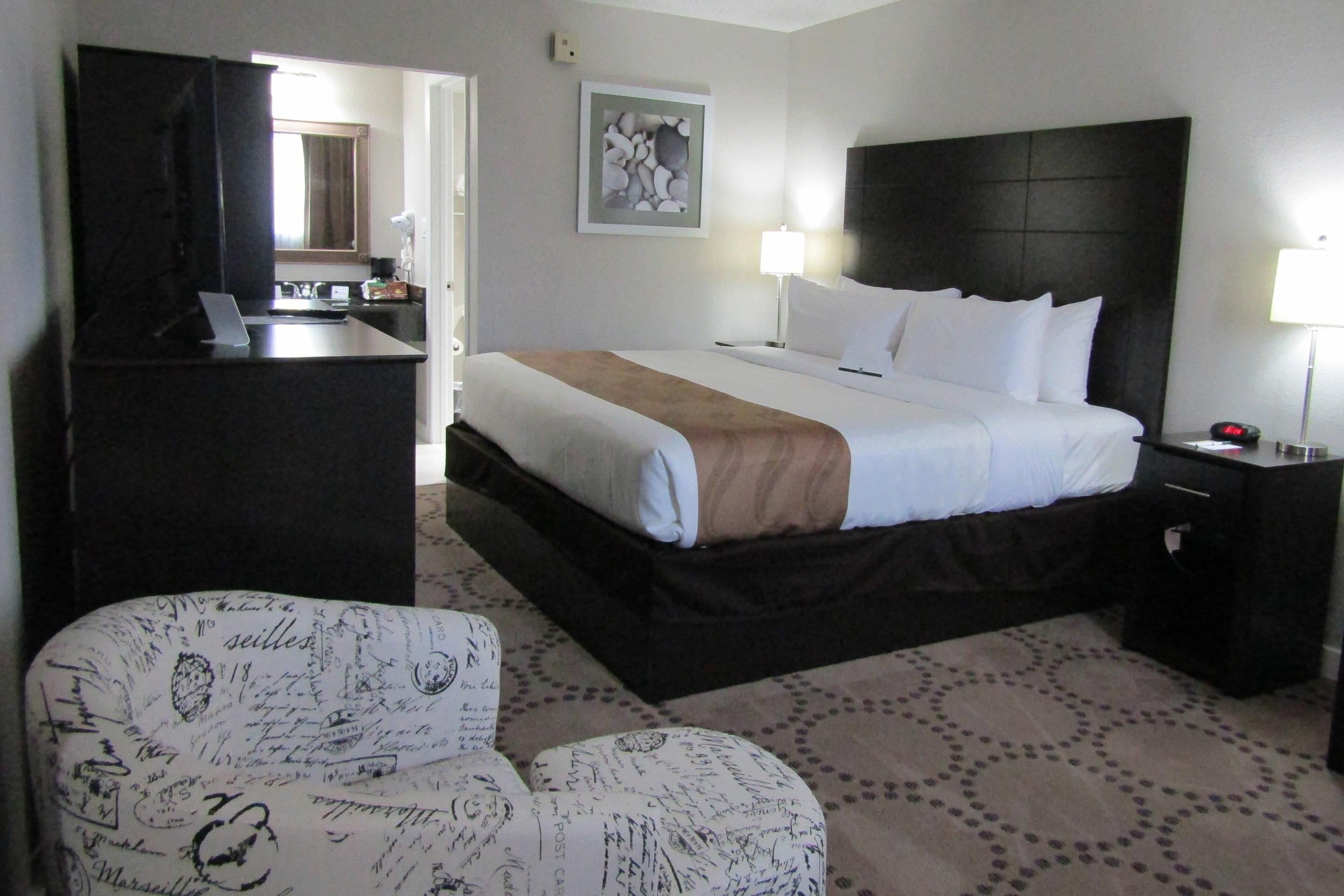 Quarto de hotel com cama, mesa de cabeceira, luminária e uma cadeira com almofada estampada.