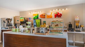 Un moderno vestíbulo de hotel con un mostrador central, máquinas de café, dispensador de jugo fresco y una exhibición de pan y pastelería.
