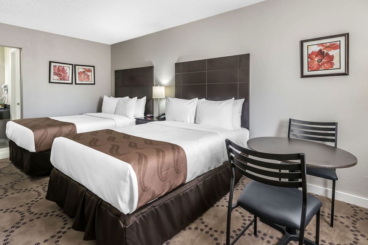 A hotel room with two beds, a desk, a chair, and a lamp.