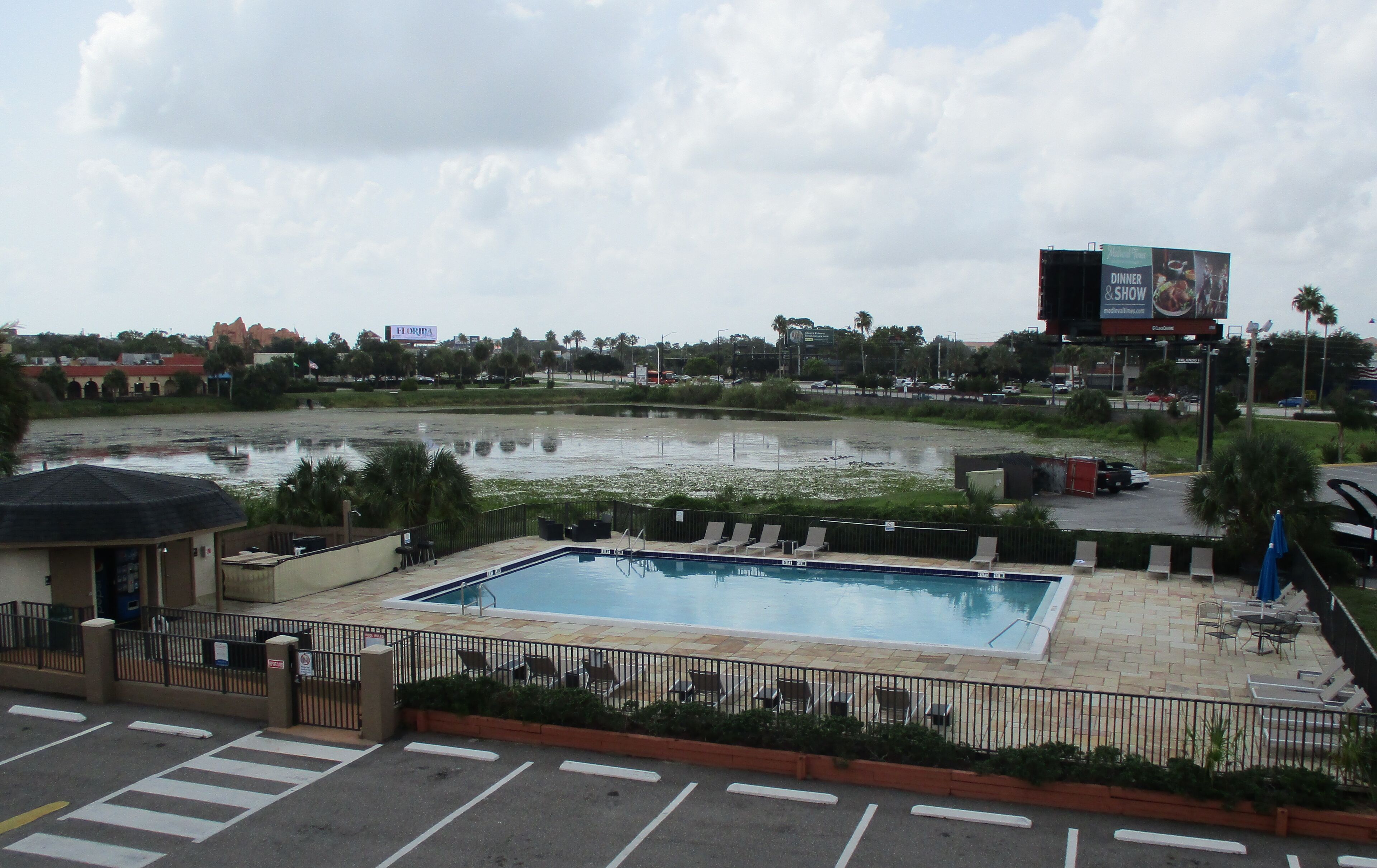 A swimming pool with lounge chairs, a fenced area, and a view of a cityscape with buildings and a billboard.