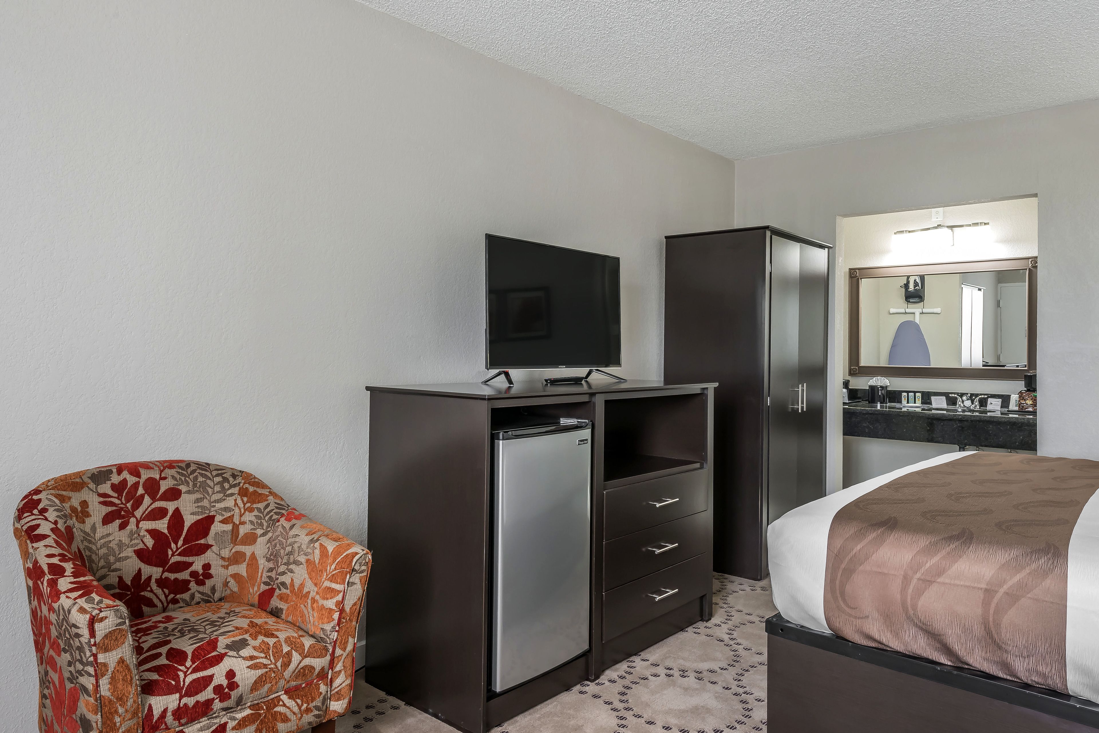 A hotel room with a bed, a television on a dresser, a chair, and a refrigerator.