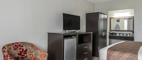 A hotel room with a bed, a television on a dresser, a chair, and a refrigerator.
