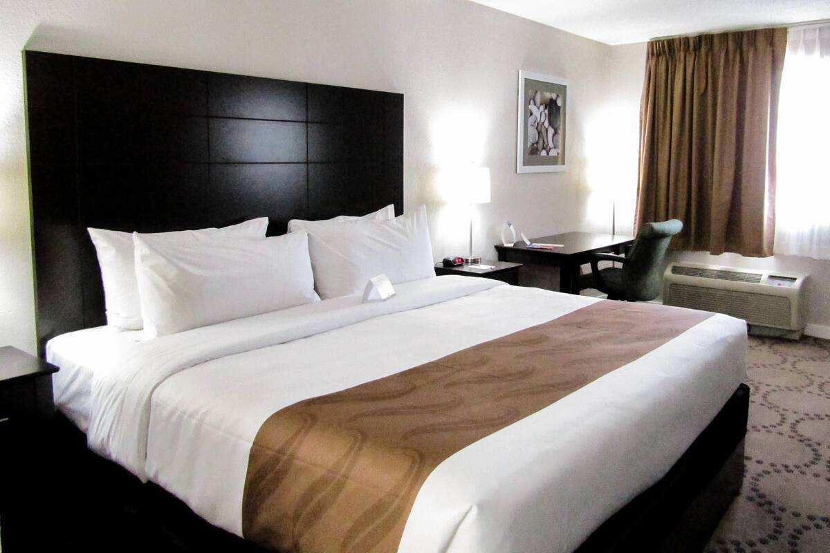 A hotel room with a large bed, a desk, a chair, and an air conditioning unit.