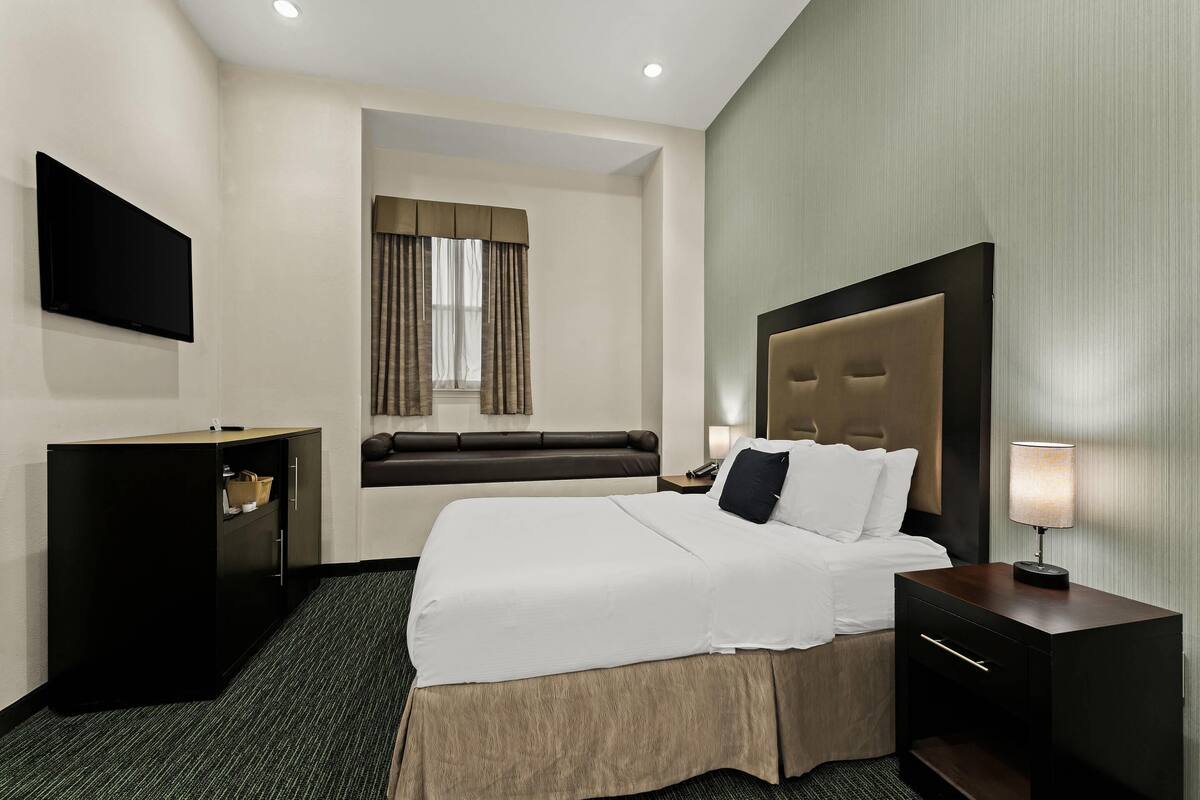 A hotel room with a large bed, a bedside table with a lamp, a TV mounted on the wall, and a window with curtains.
