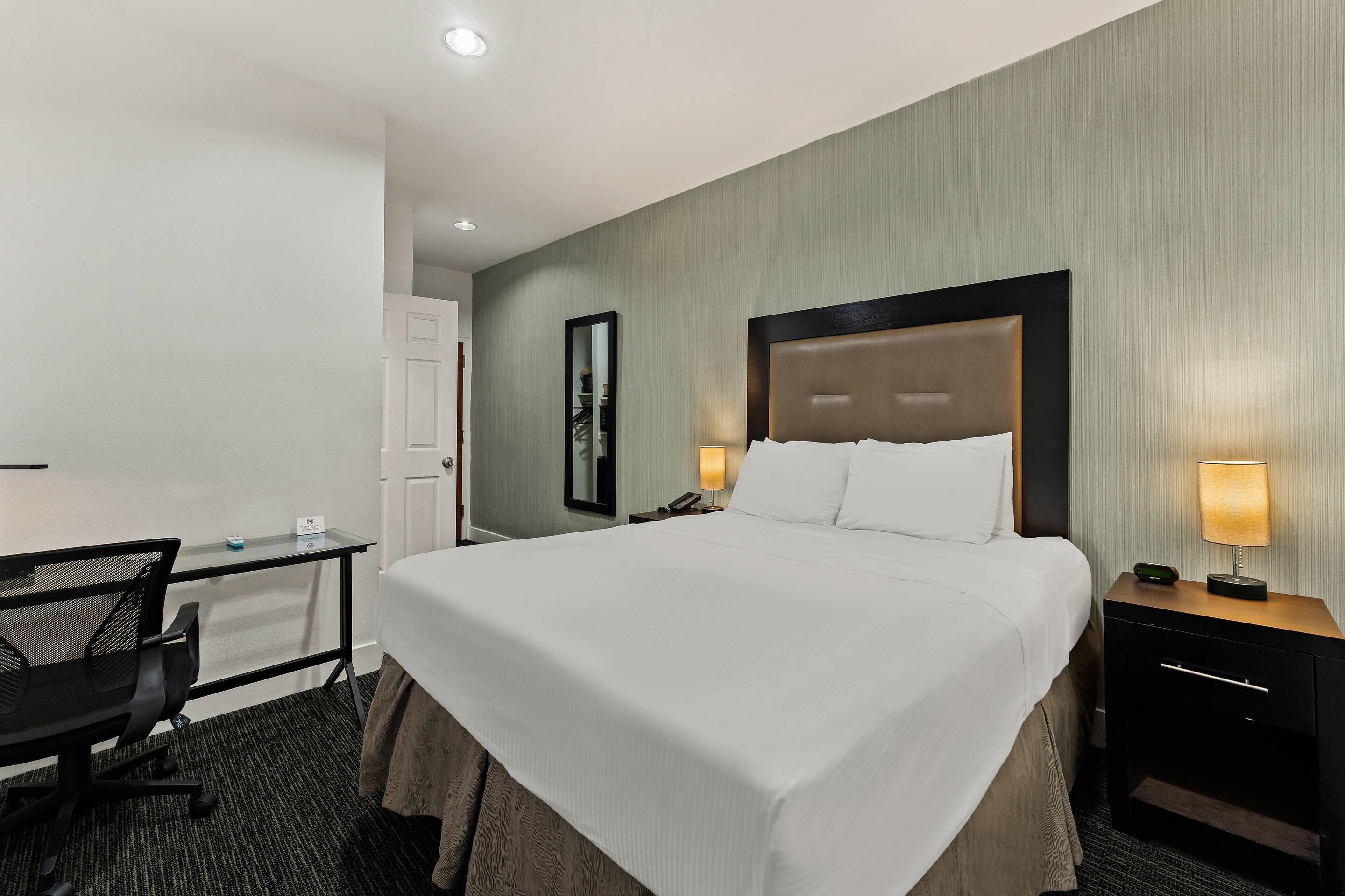 A hotel room with a large bed, a desk, a chair, a nightstand, and a mirror.