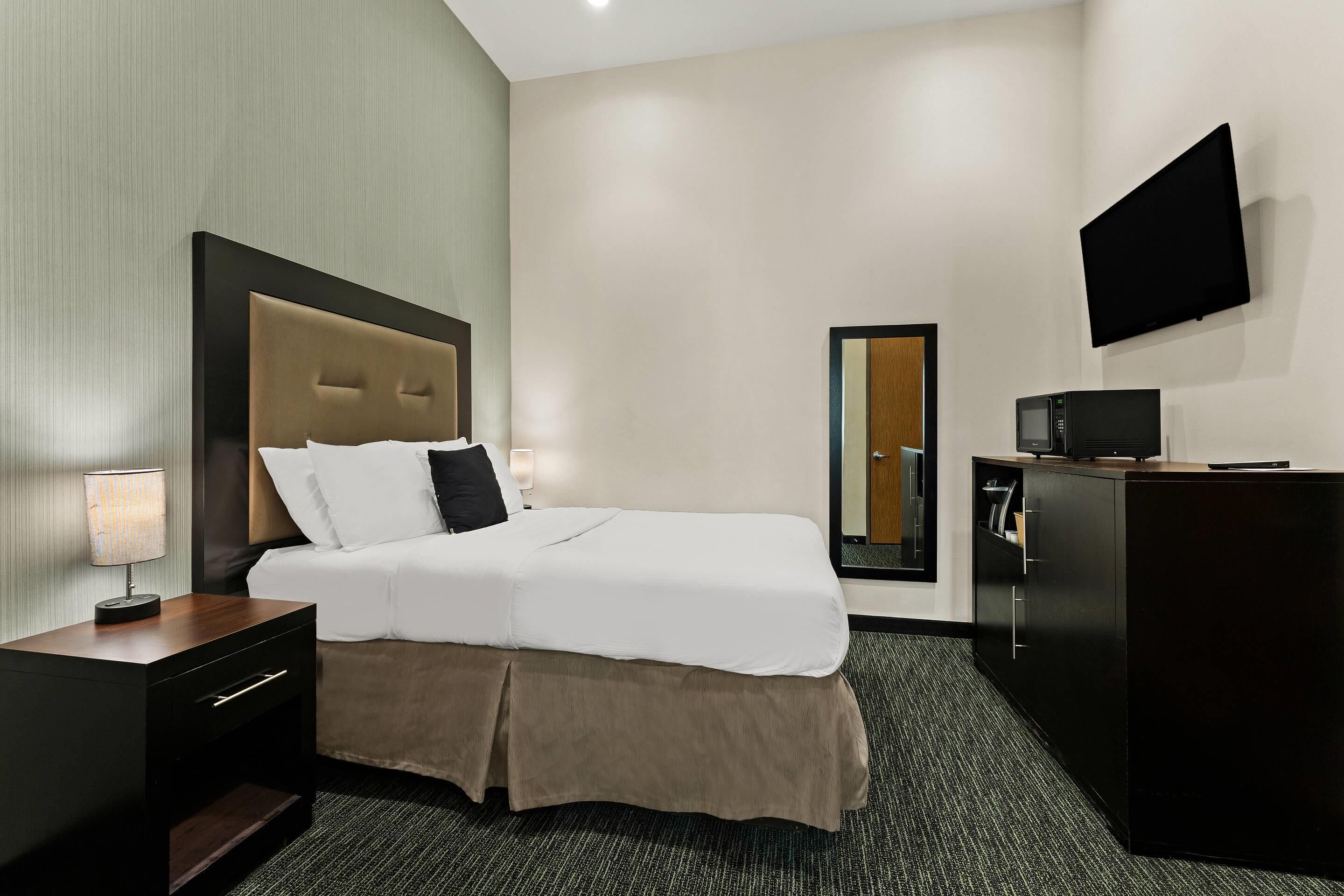 Una habitación de hotel con cama, mesita de noche, lámpara, televisor y espejo.