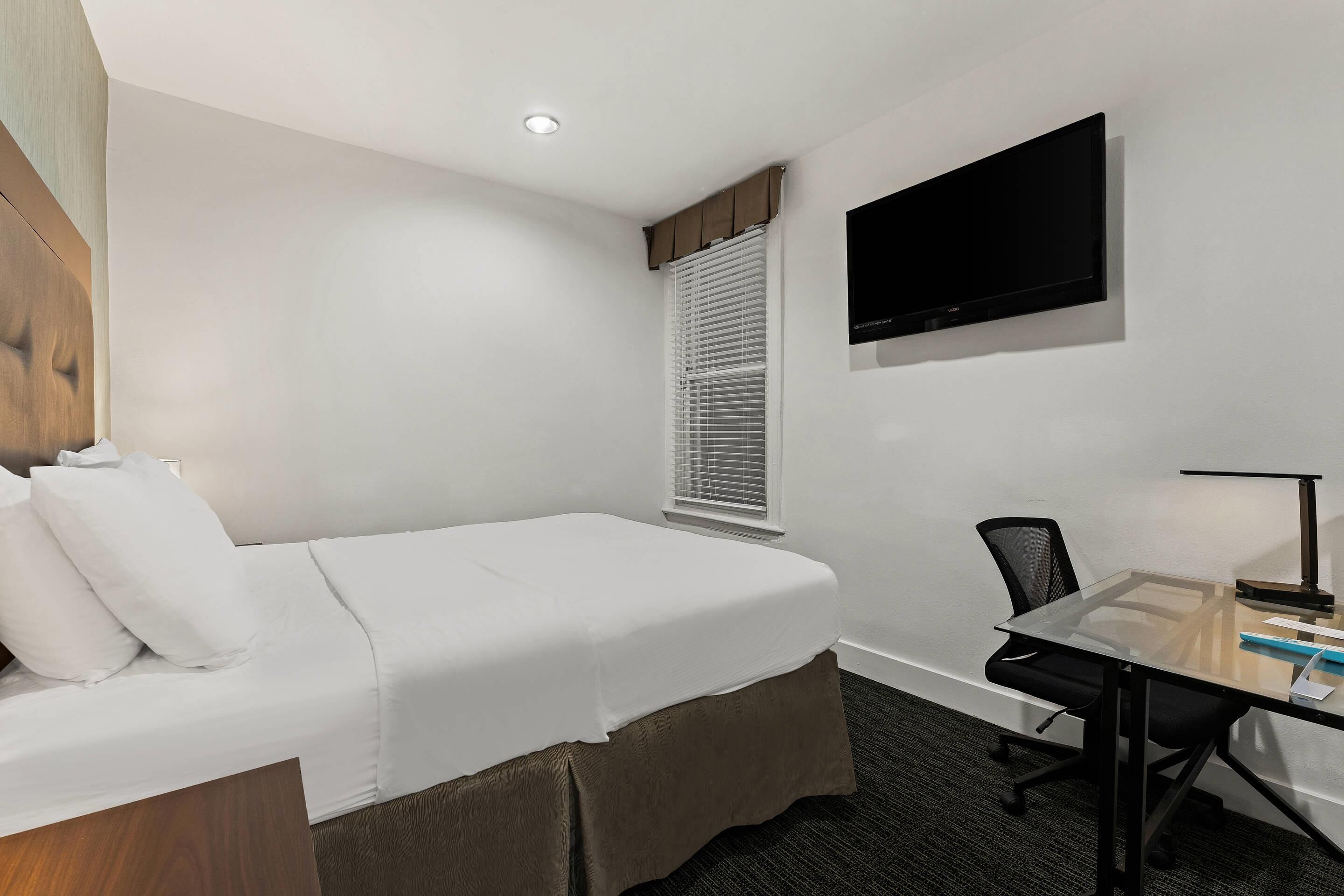 Habitación de hotel con una cama individual, un escritorio con silla, un televisor de pantalla plana en la pared y una ventana con persianas.